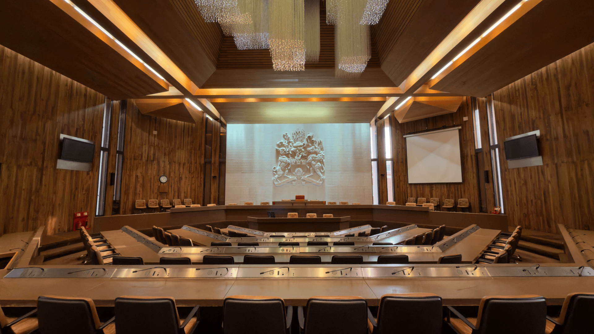 Large council chamber with tiered seating arranged in a horseshoe shape, wooden panelled walls, and a high ceiling featuring modern light fixtures.